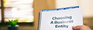 A hand holds a spiral notebook titled "Choosing A Business Entity," with divider tabs labeled "Partnership," "Corp," "S Corp," and "LLC" visible at the top.