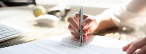 A person’s hand holding a pen and writing on a printed document on a desk near a computer keyboard and mouse.