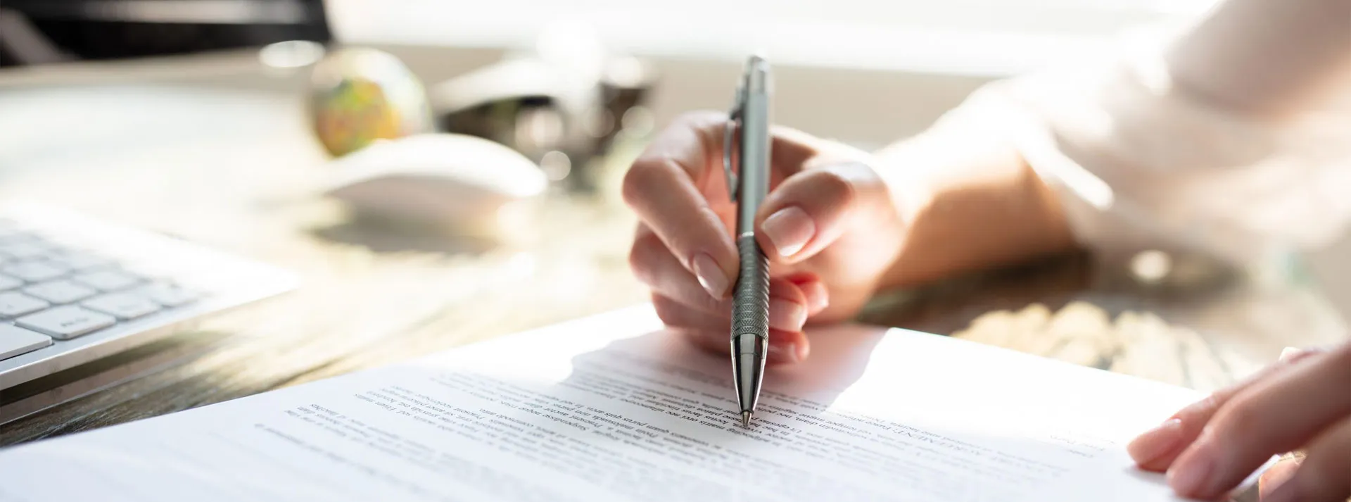 A person’s hand holding a pen and writing on a printed document on a desk near a computer keyboard and mouse.