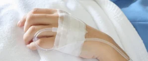 Close-up of a hand with an intravenous (IV) line inserted and secured with medical tape, resting on a white blanket.