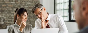 Two business professionals sit at a desk, one whispering to the other while looking at a third person holding documents in the foreground.