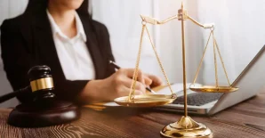 A person in business attire works on a laptop at a desk with a gavel and a golden balance scale in the foreground.
