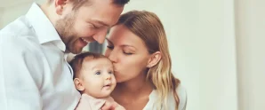 A man and woman hold and kiss a baby indoors, all three facing the camera with gentle expressions.