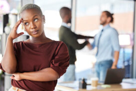 A woman stands with her hand on her head, looking away thoughtfully—perhaps reflecting on gender discrimination at Berkeley—as two people converse in the blurred background of an office setting.