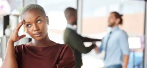 A woman stands in the foreground with a thoughtful expression while two people have a conversation in the blurred background of an office setting.