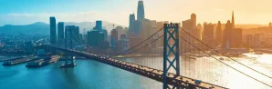 Bay Bridge spans across water with cars in both directions, leading toward the San Francisco skyline under a partly sunny sky.